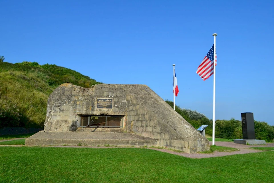 A concrete bunker with French and American flags flying from flagpoles, situated on a grassy area beneath a clear blue sky.