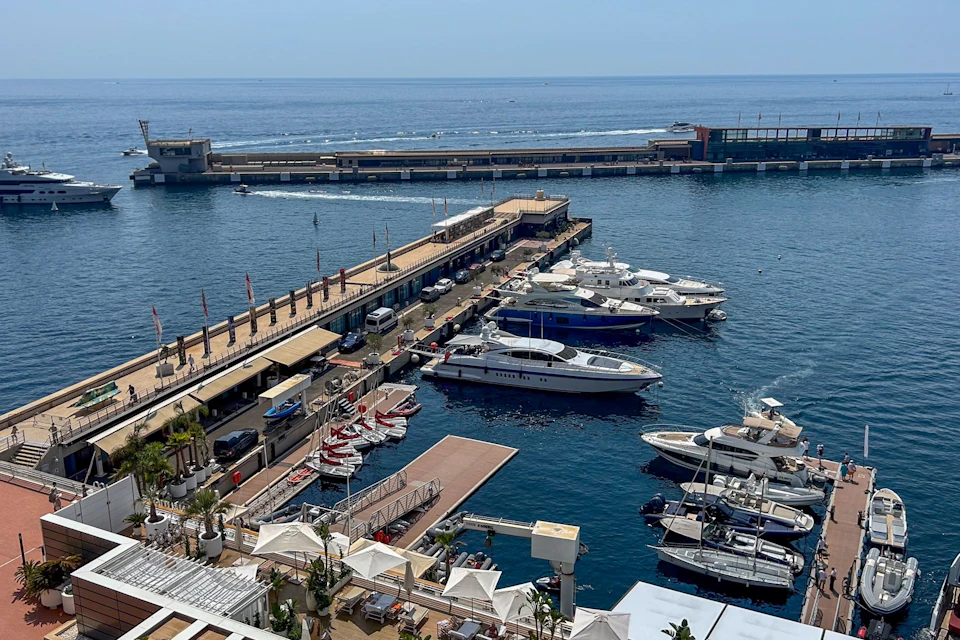 A harbor view featuring luxury yachts docked along piers, with a promenade and the sea in the background under a clear sky.