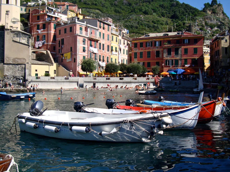 Colorful boats docked at a harbor with people swimming and sunbathing. Background includes colorful buildings and a hillside.