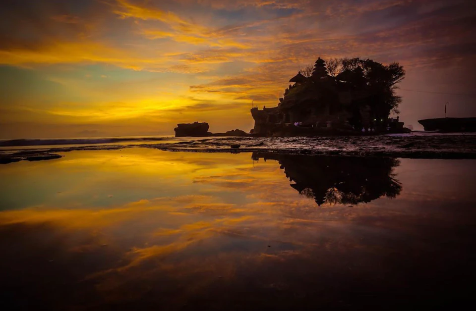 Sunset over a rocky coastline with a temple silhouetted against the sky, reflecting in a calm body of water.