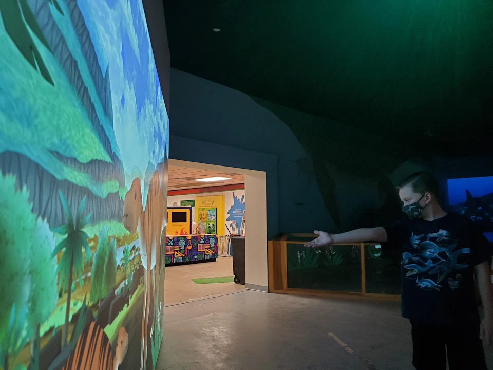 A child wearing a mask interacts with a large, colorful wall projection of a prehistoric landscape inside a building.