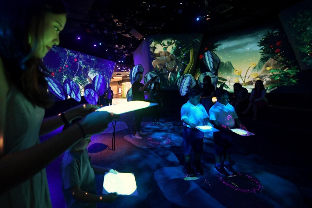 Lidé v temné místnosti držící zářící tablety pod modrým a fialovým okolním světlem s přírodními scénami promítanými na stěny.