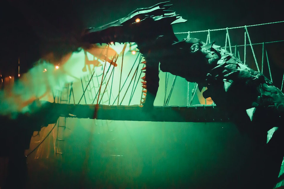 A large, mechanical dragon with glowing eyes on a misty suspension bridge, illuminated by green and orange lighting.
