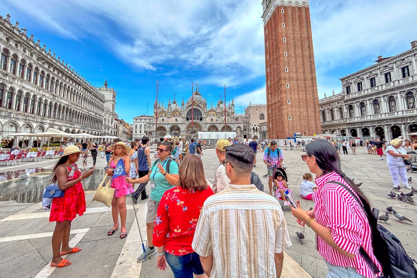 Ein belebter Platz mit Menschen, die sich unter freiem Himmel in der Nähe historischer Gebäude versammeln, darunter ein hoher Glockenturm und eine verschnörkelte Fassade.