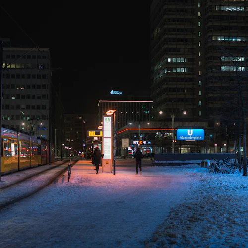 Stadsgata på natten med spårvagn, snötäckt mark, människor som går, neonljus och höga byggnader. På skylten står det "Alexanderplatz".