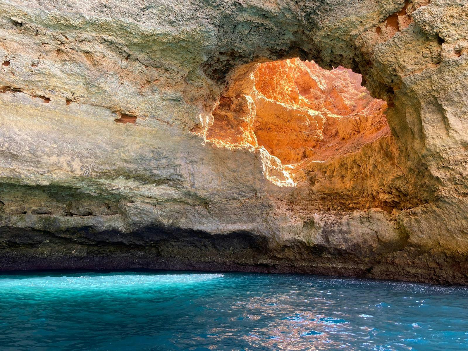 Bright sunlight shines through a rocky cave opening, illuminating orange-brown rock formations above blue water below.