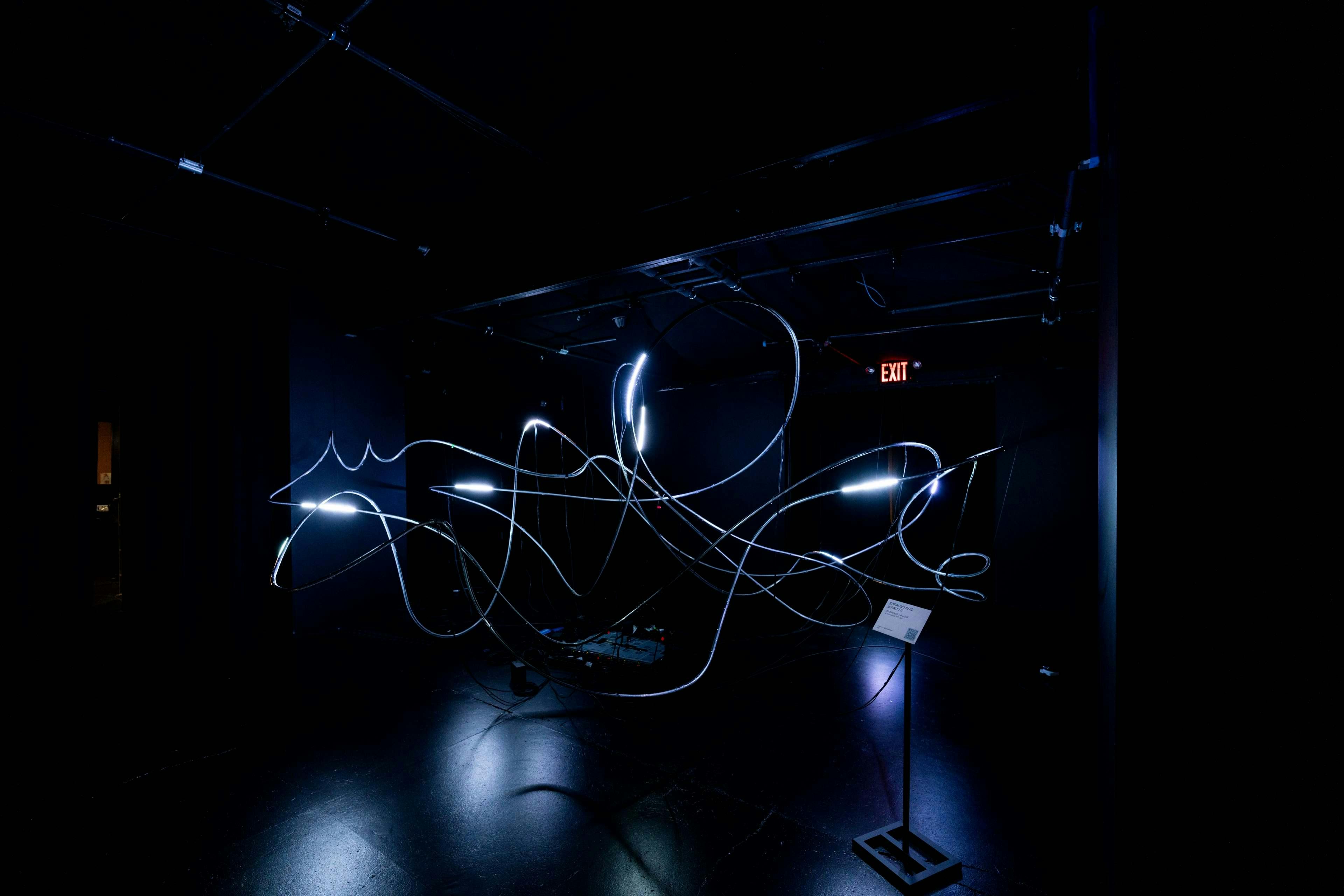 Dunkler Raum mit beleuchteten, sich drehenden Lichtschläuchen und einem "EXIT"-Schild im Hintergrund.