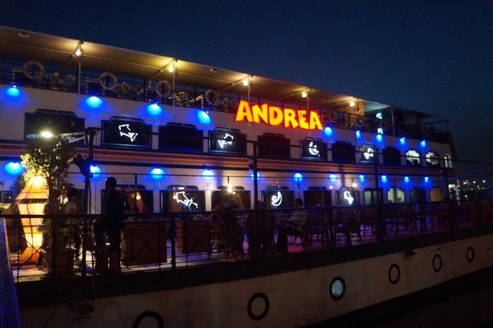 A brightly lit boat named Andrea with blue and white lights, featuring a deck with tables and chairs, docked at night.