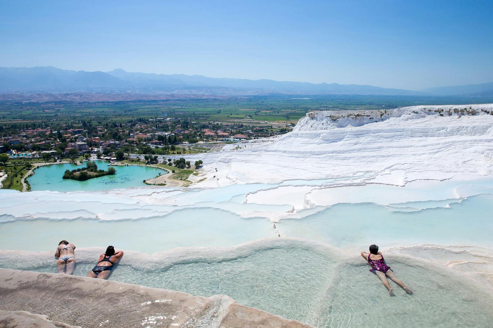 Two people relax in natural terraces filled with turquoise water, overlooking a landscape with mountains and a town in the distance.