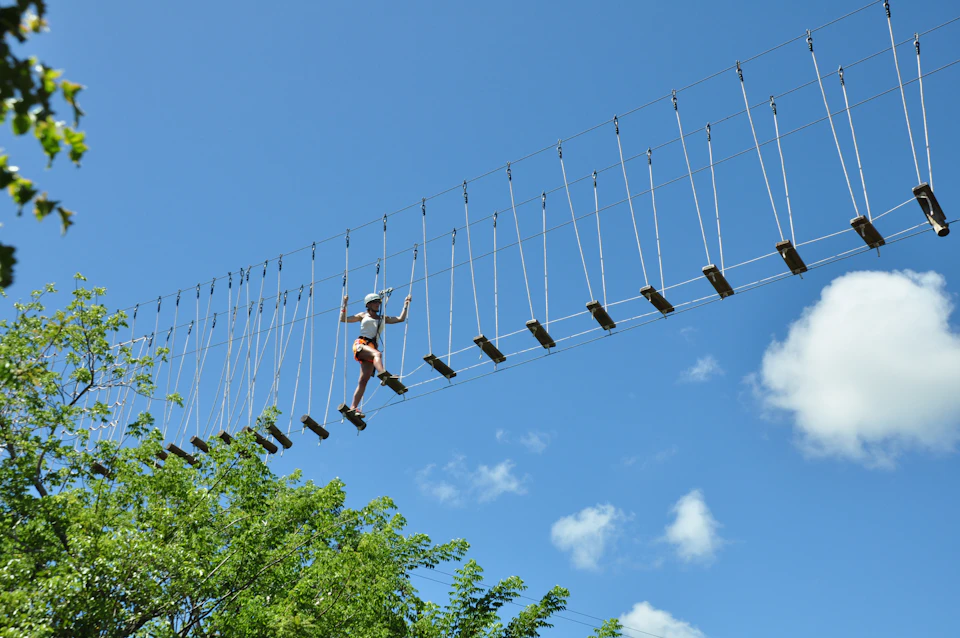 A person wearing safety gear navigates a suspended rope bridge high above the ground against a clear blue sky. Trees are visible below.