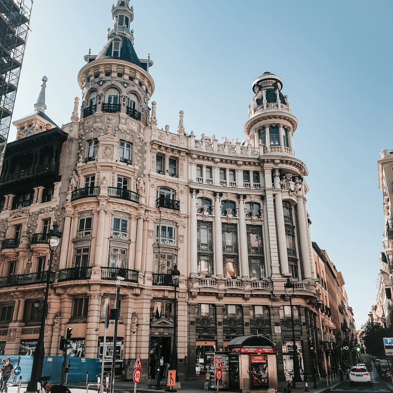 Utsmyckad historisk byggnad med höga spiror en solig dag. Stadsgata med fotgängare, trafikskyltar och byggbarriärer.