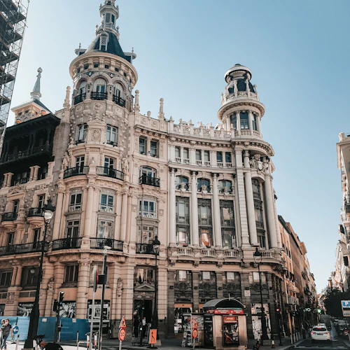 Utsmyckad historisk byggnad med höga spiror en solig dag. Stadsgata med fotgängare, trafikskyltar och byggbarriärer.