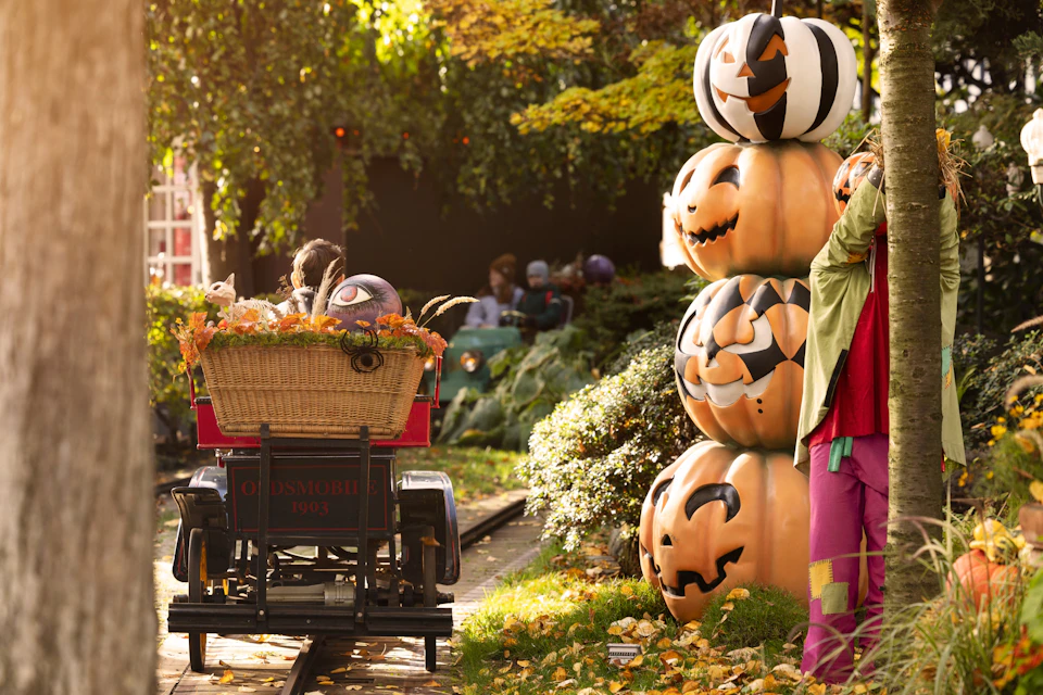 People riding in a vintage-themed cart with a basket of fall decor, passing a stack of carved pumpkins in a park-like setting.