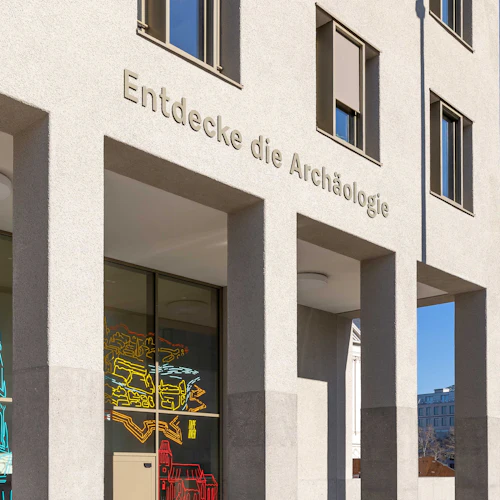 Byggnadens fasad med "Entdecke die Archäologie" inskription, färgglada neonskyltar och TV-torn i bakgrunden.