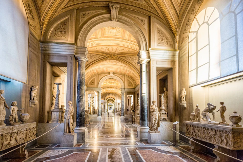 An ornate corridor with archways, marble statues, and a decorated ceiling, illuminated by natural light from tall windows.