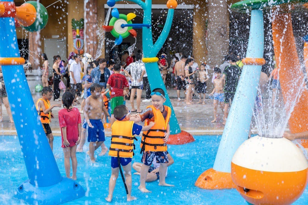 Children playing in a splash zone with water features, wearing life vests, surrounded by other kids and adults in swimwear.