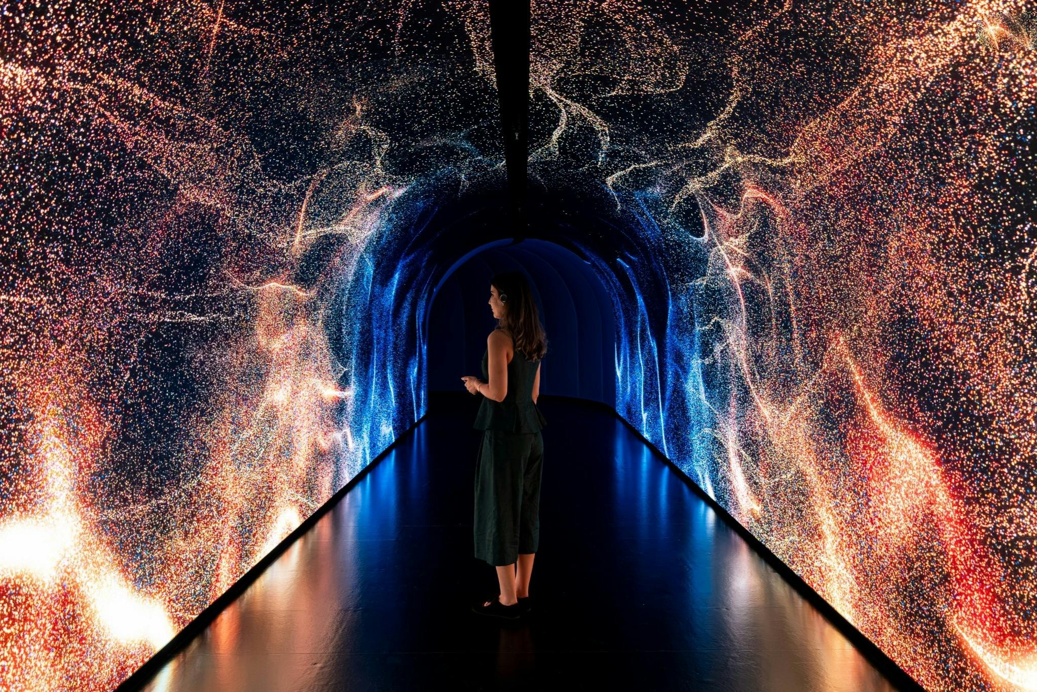 A person stands in a tunnel illuminated by dynamic blue and orange light patterns on the walls, creating an immersive effect.