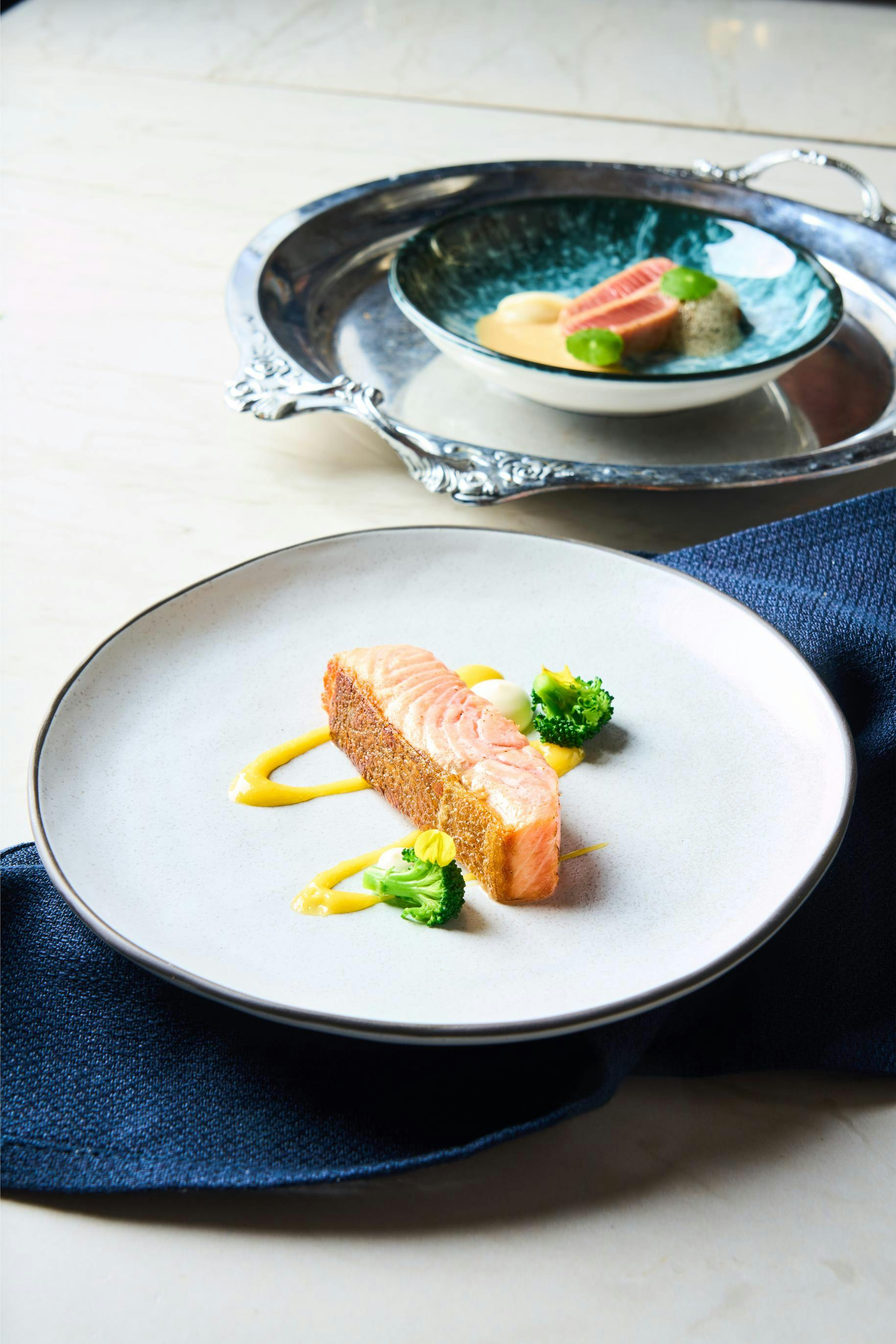 A plated dish featuring a seared salmon piece with sauce and broccoli on a white plate, with a second dish in the background.