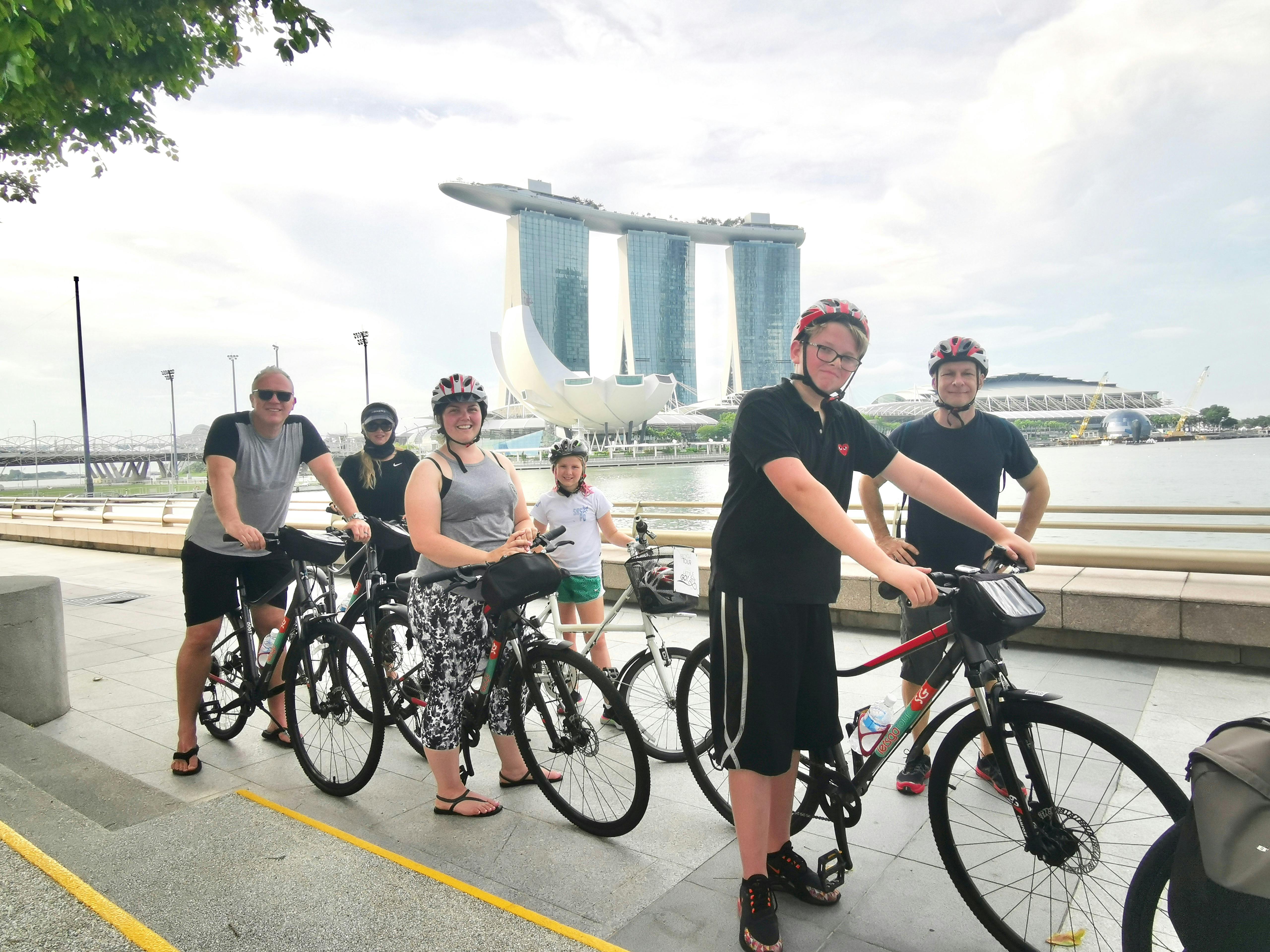 En gruppe cyklister poserer med deres cykler langs en havnefront med Marina Bay Sands i baggrunden under en delvist overskyet himmel.