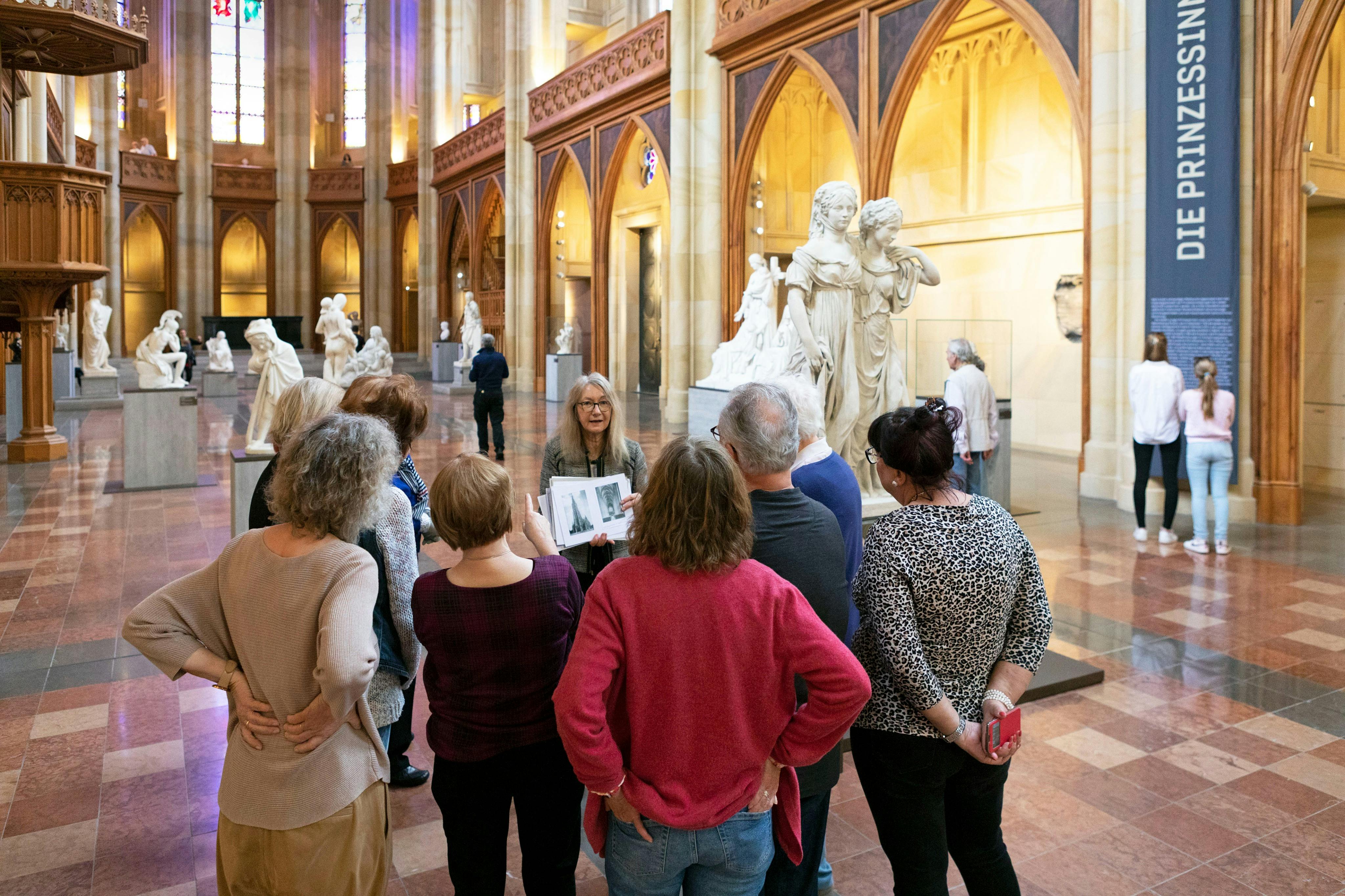 En grupp människor i en museumshall som observerar skulpturer och lyssnar på en guide som håller i en bok med bilder.