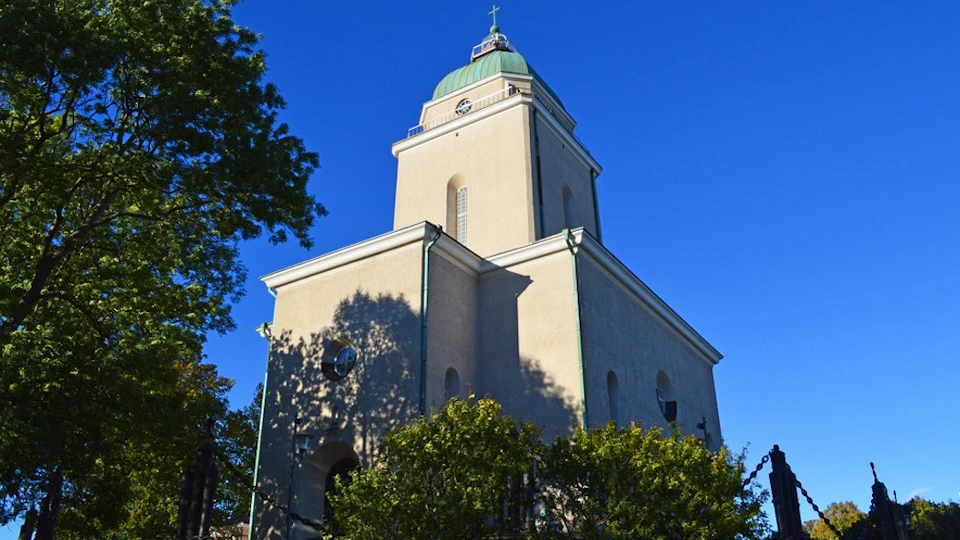 A tall beige church with a green dome and cross on top, surrounded by trees, under a bright blue sky.