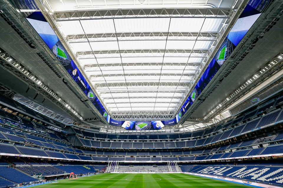 A large stadium with a retractable roof, LED screens, and a green football field, viewed from the goal area.