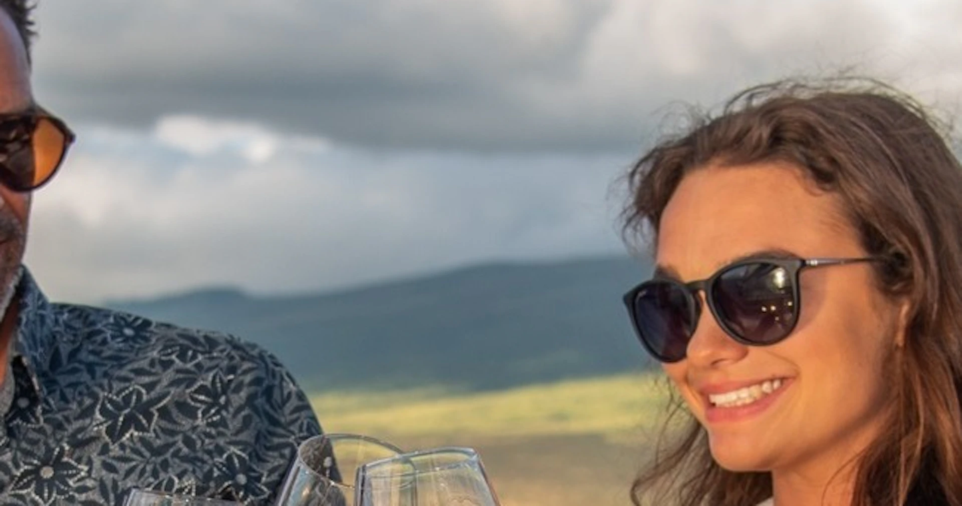 Three people in sunglasses toast with glasses of white wine on a boat, with water and hills in the background.