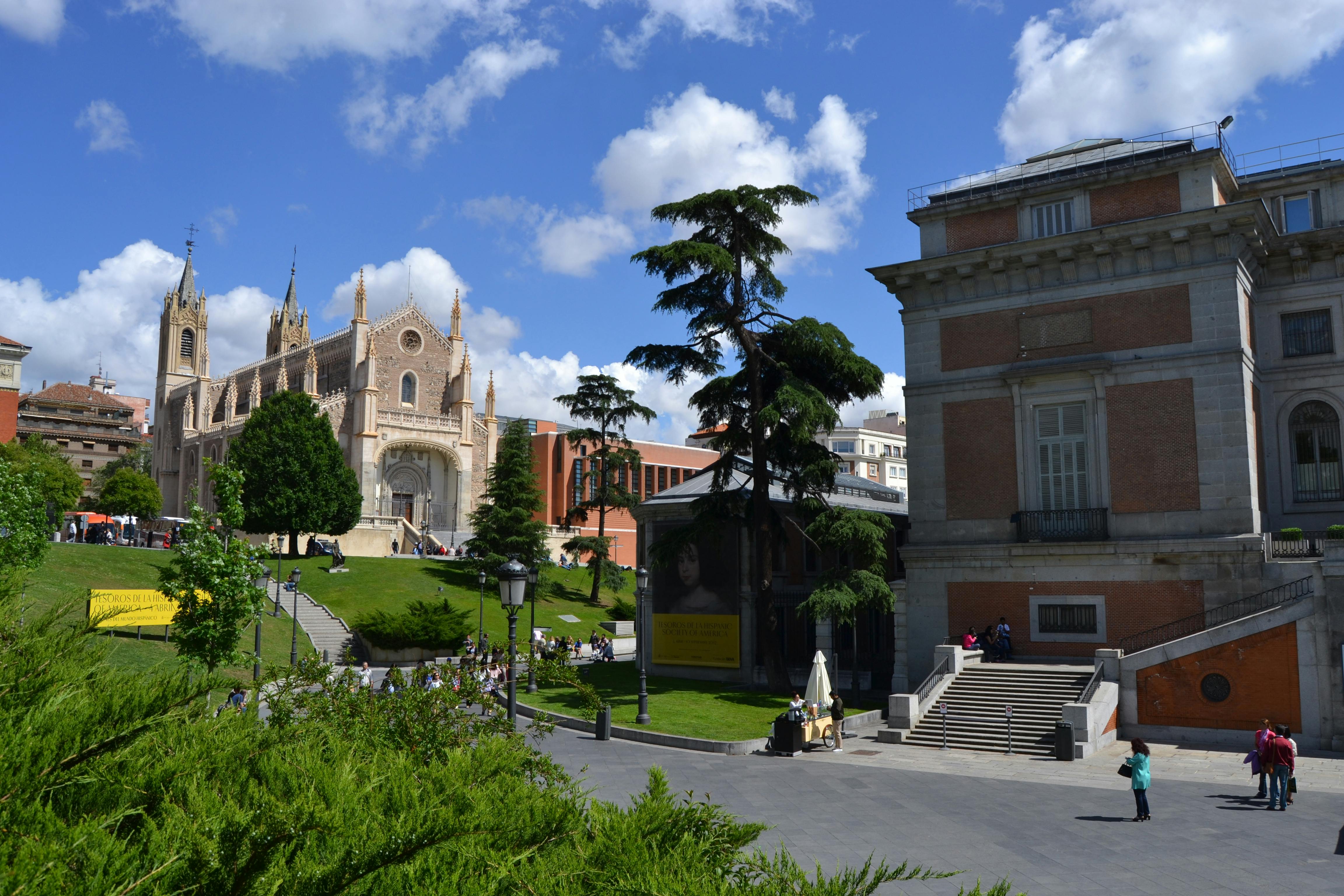 Eine historische Kirche, ein Baum und ein Museumsgebäude mit Treppen auf einem Platz unter einem teilweise bewölkten blauen Himmel.