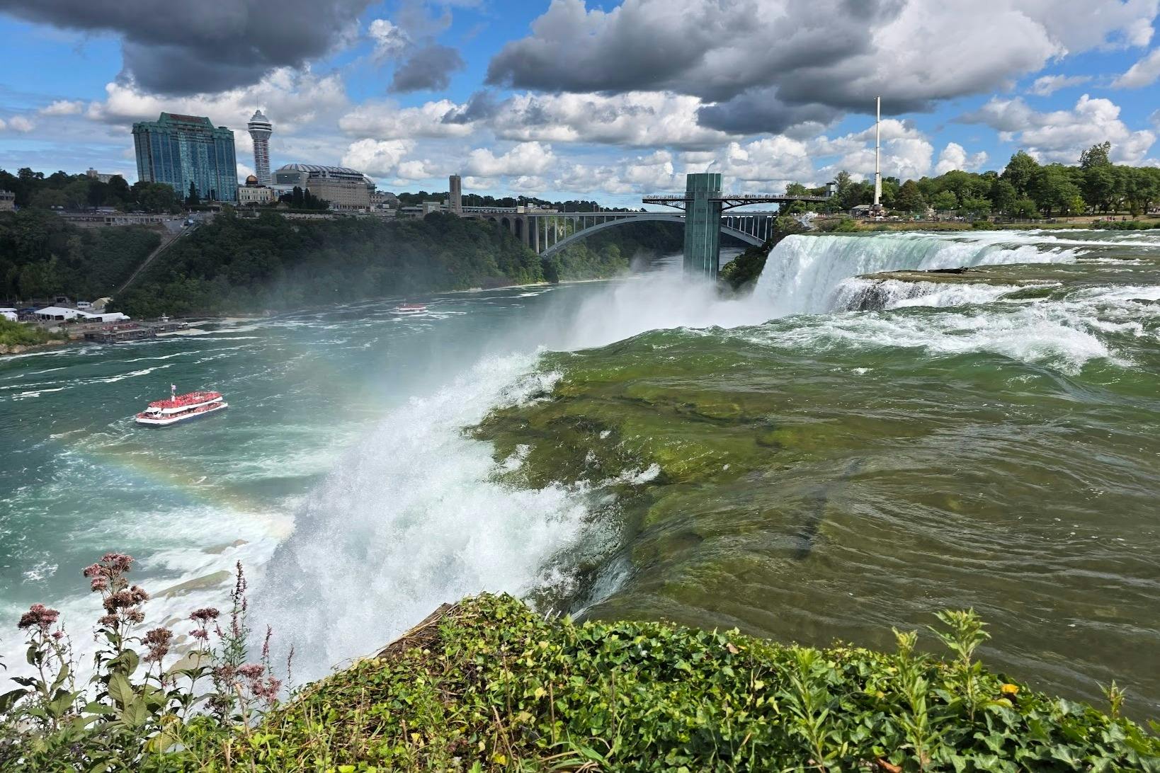 俯瞰尼亚加拉大瀑布（ Niagara Falls ） ，这里有瀑布般的水流、郁郁葱葱的绿色植物、彩虹，背景是多云的天空下的城市景观。