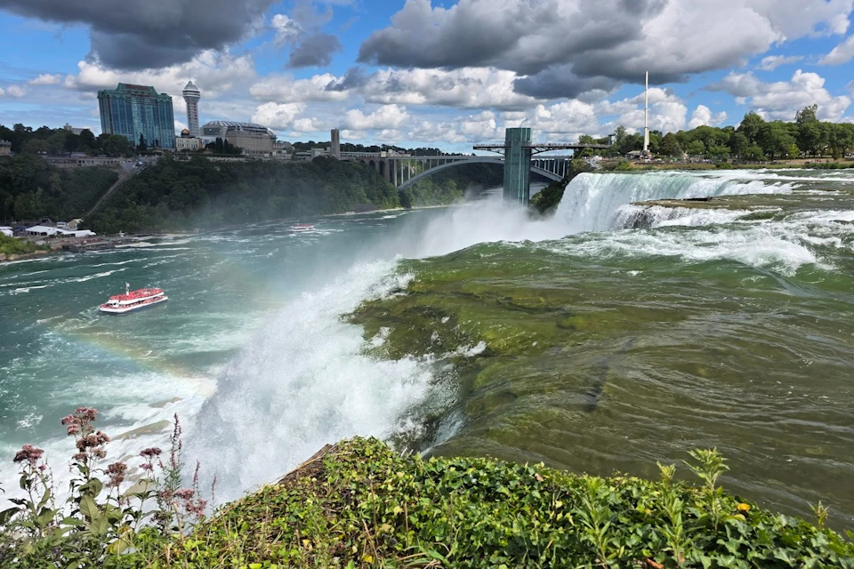 A view of Niagara Falls with cascading water, lush greenery, a rainbow, and the cityscape in the background under a cloudy sky.