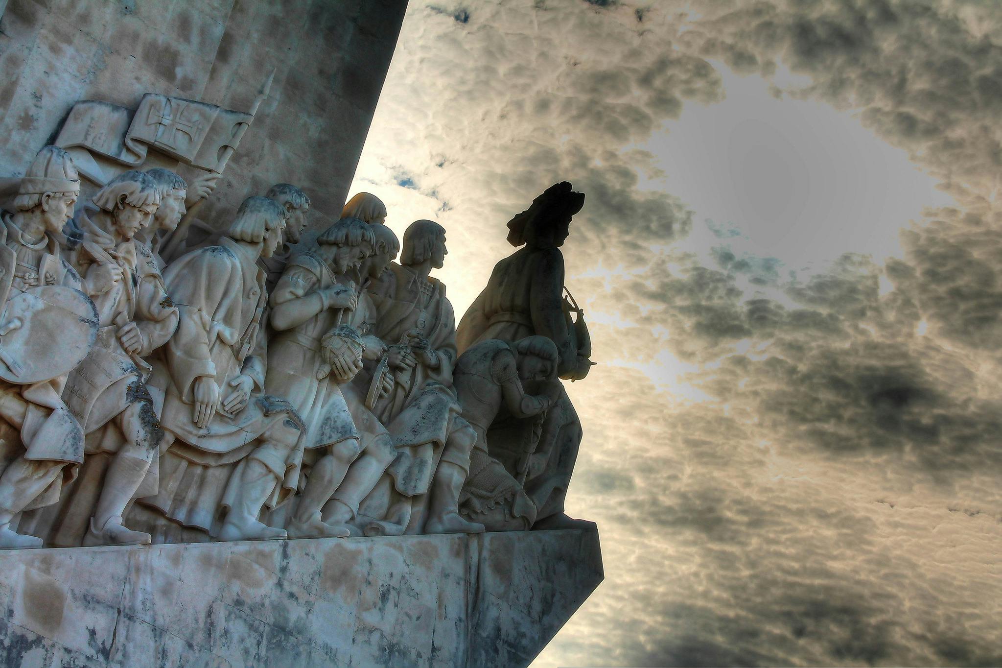 Silhuettade stenstatyer av historiska figurer i rad mot en molnig, bakgrundsbelyst himmel.