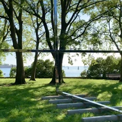 En gräsbevuxen park med träd, en polerad tvärstruktur i metall och utsikt över en sjö med segelbåtar i bakgrunden.