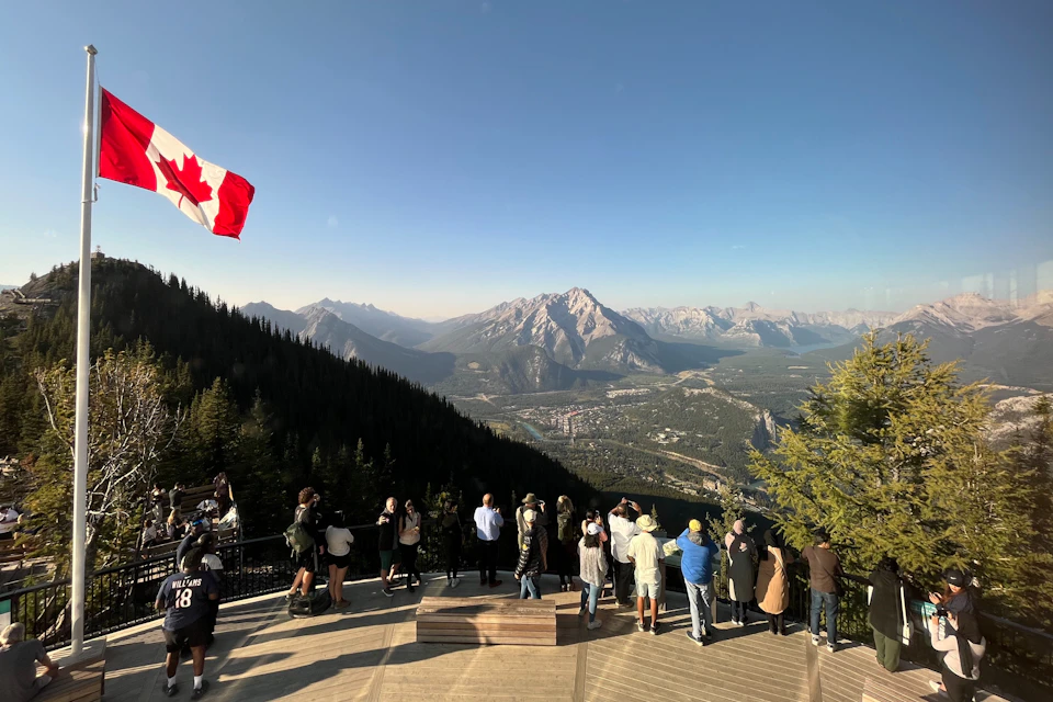 People stand on a viewing platform overlooking a vast mountainous landscape with a Canadian flag flying.