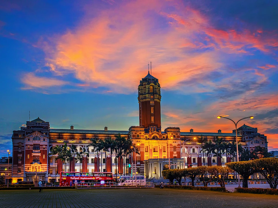 A historic building with a tall central tower under a colorful sunset sky, surrounded by palm trees and streetlights.