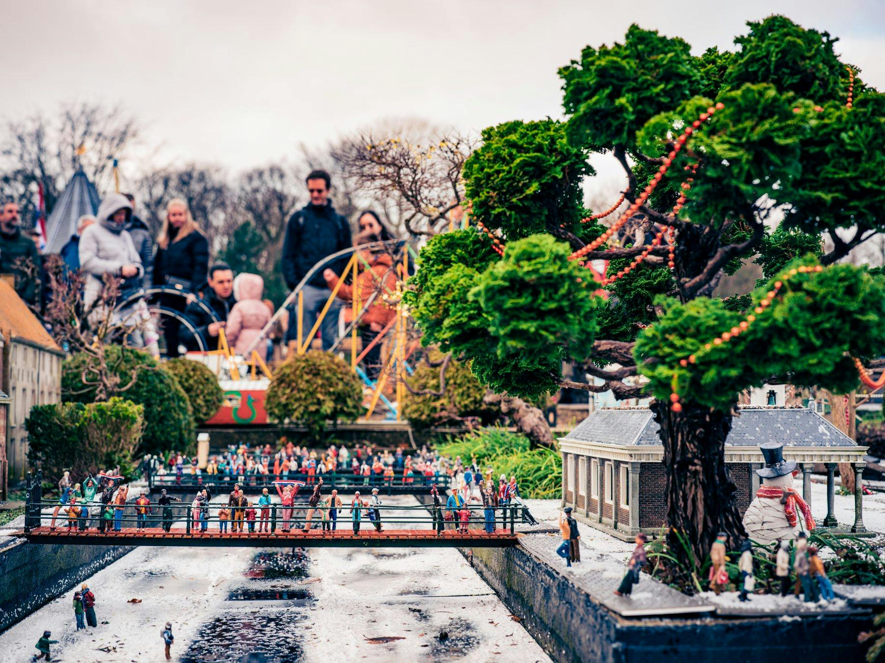 ミニチュアシーンでは、モデルの人々が運河の橋の上に立っており、木々や建物に囲まれています。背景には本物の人々がぼやけています。