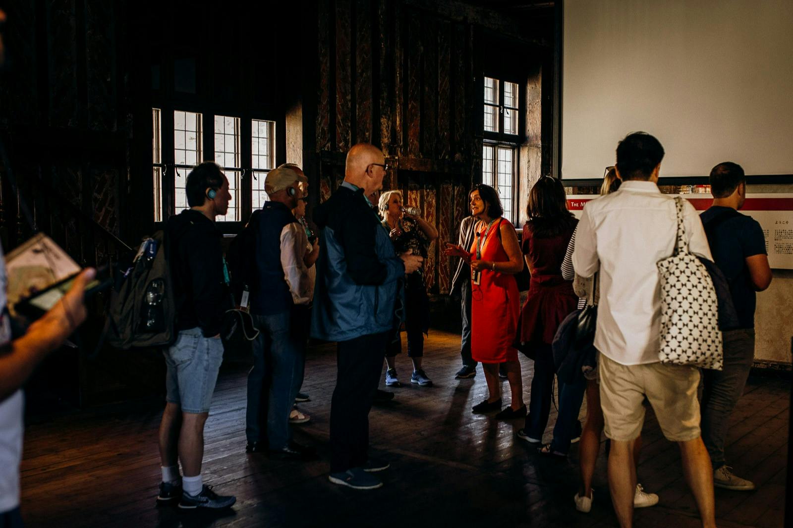 A group of people attentively listening to a presenter in a dimly lit room with wooden floors and large windows.