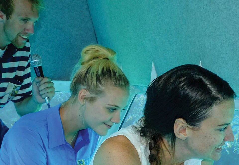 Three people inside a submarine look out of windows at underwater coral. The woman in front points with a smile.