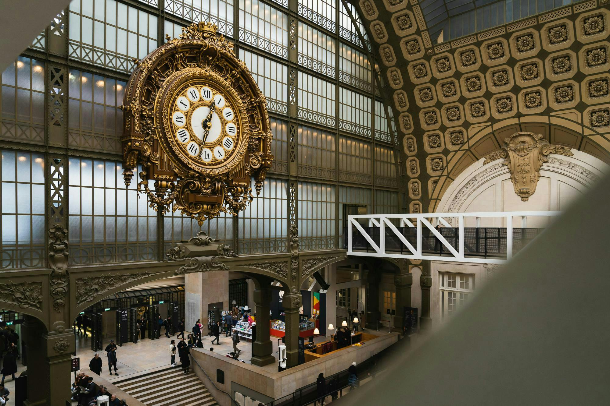 커다란 기차역 안에는 화려한 황금색 시계가 걸려 있으며, 높은 천장과 아치형 창문, 아래로 걸어가는 사람들이 있습니다.