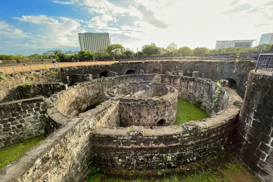 A historic stone fort with circular structures and grass in the center, surrounded by urban buildings and greenery.