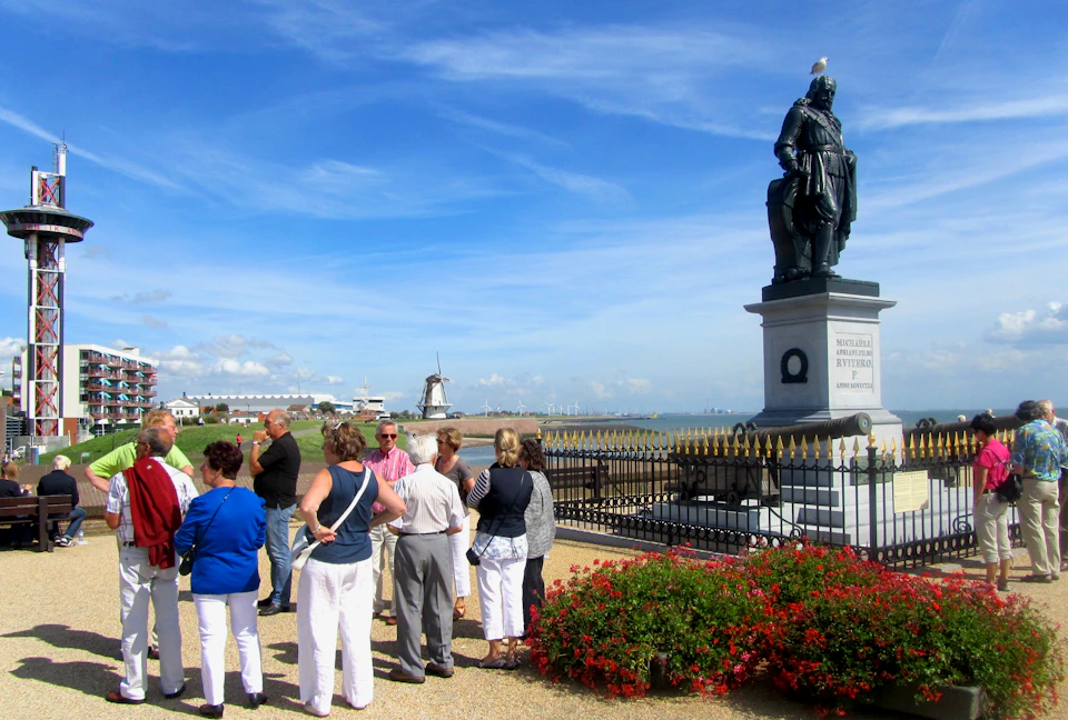A group of people stand near a statue with a bird on top, surrounded by a fence. Windmill and coastline visible in the background.