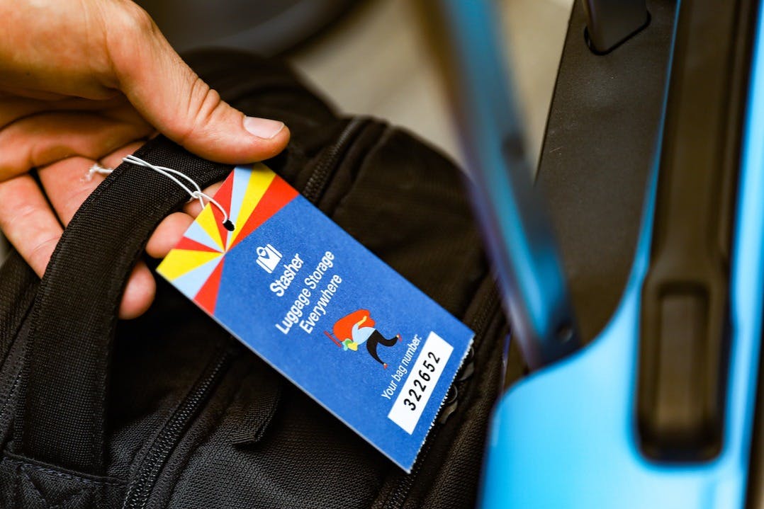 Ruka držící černou tašku s barevným štítkem „Stasher Luggage Storage Everywhere“ vedle modrého kufru.