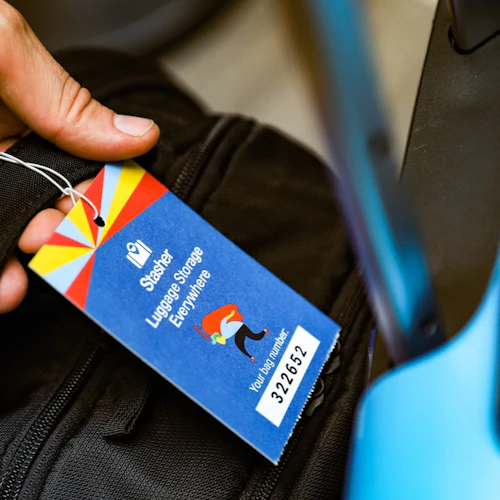 Hand som håller en svart väska med en färgglad "Stasher Luggage Storage Everywhere" -tagg, bredvid en blå resväska.