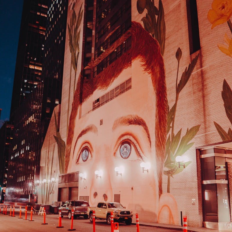 Μεγάλη τοιχογραφία του προσώπου ενός παιδιού με μπλε μάτια σε ένα κτίριο της πόλης, που περιβάλλεται από ψηλά φυτά και σταθμευμένα αυτοκίνητα κάτω τη νύχτα.