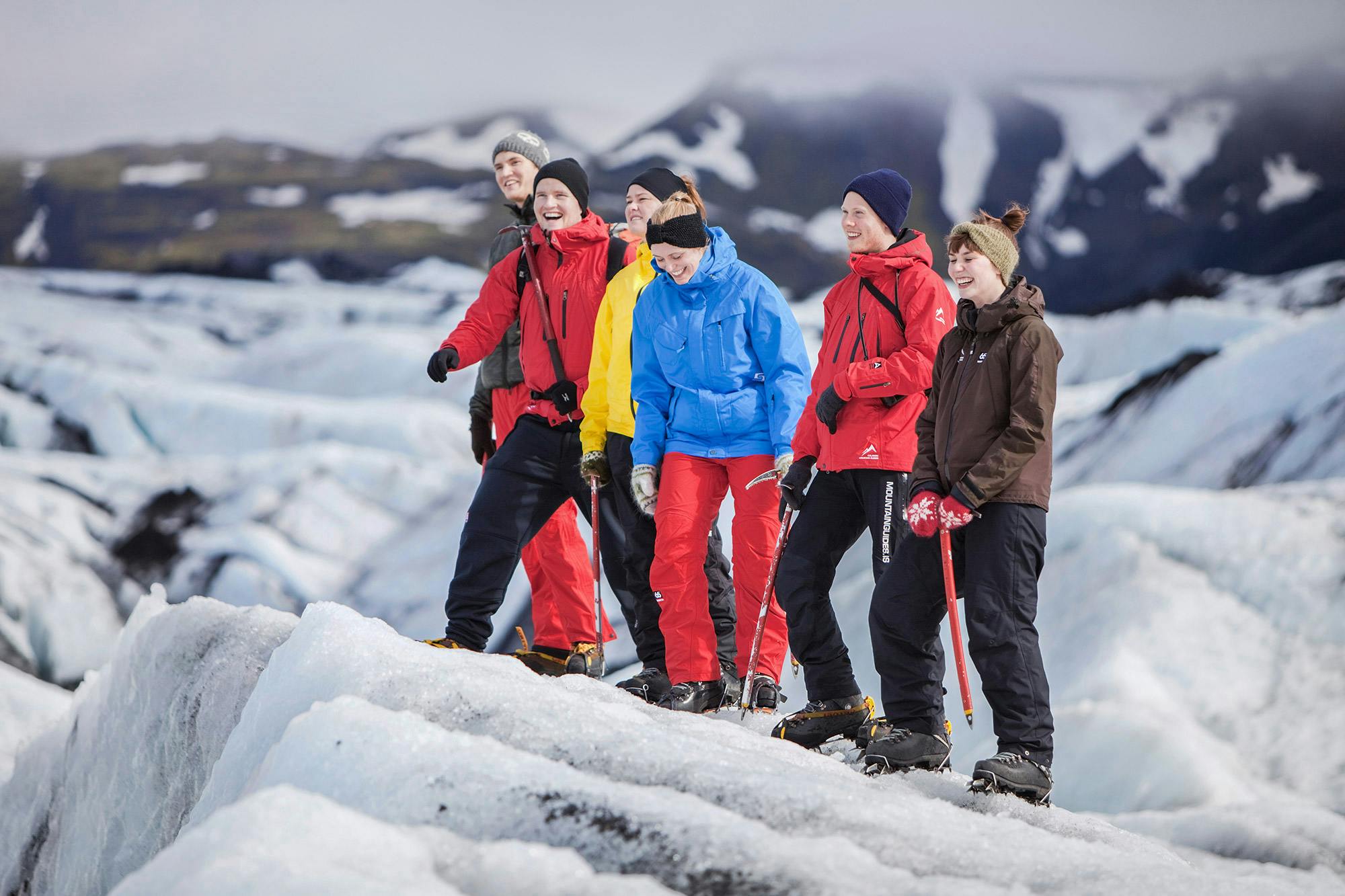 Group of people in winter clothing smiling and walking on a snowy landscape with mountains in the background.