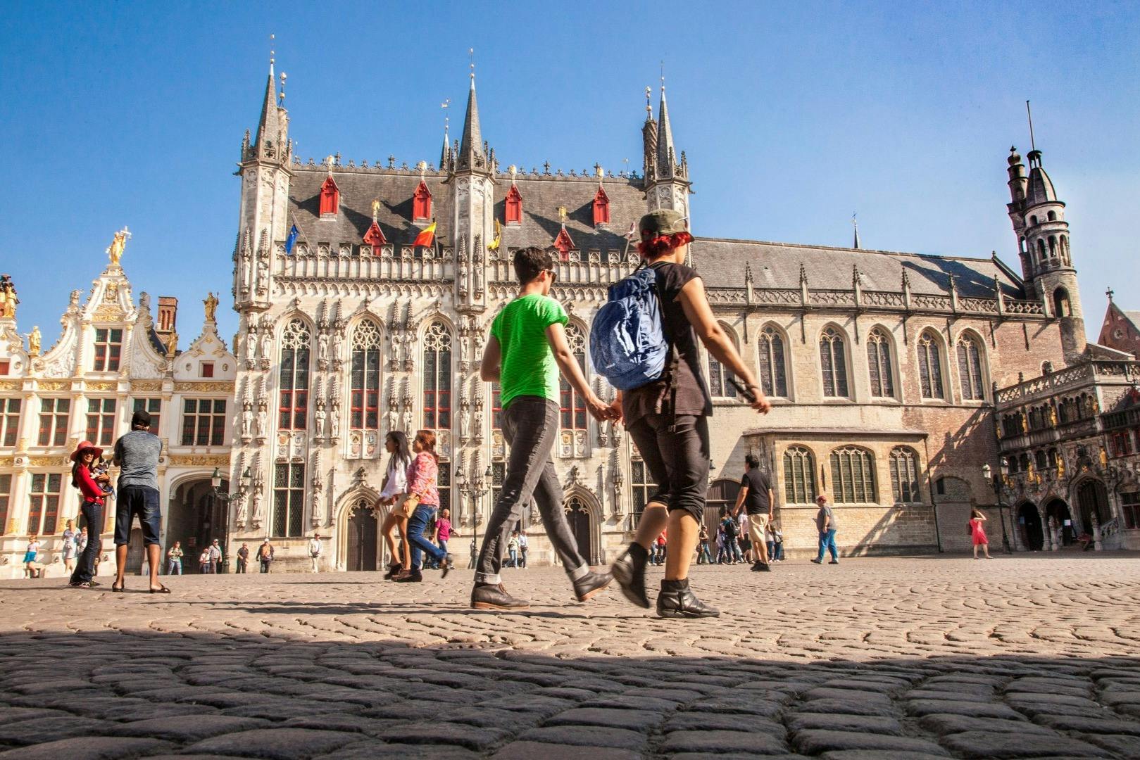 Tourists walk on a cobblestone square in front of a historic gothic-style building with spires and ornate details under a clear blue sky.