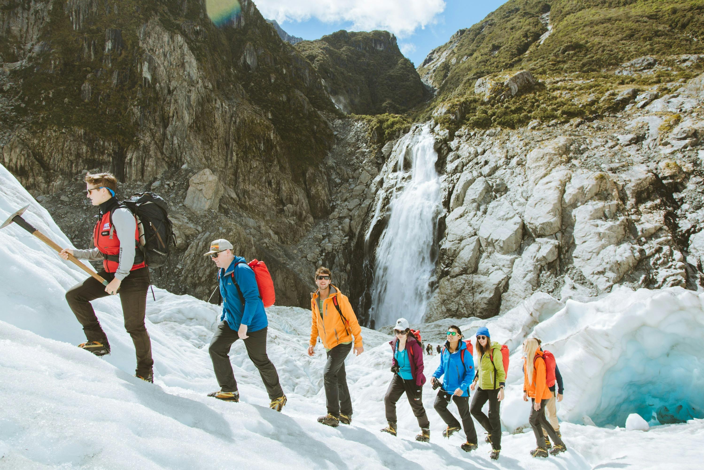 A group of five people hiking on a snowy slope with a waterfall and rocky cliffs in the background under a partly cloudy sky.