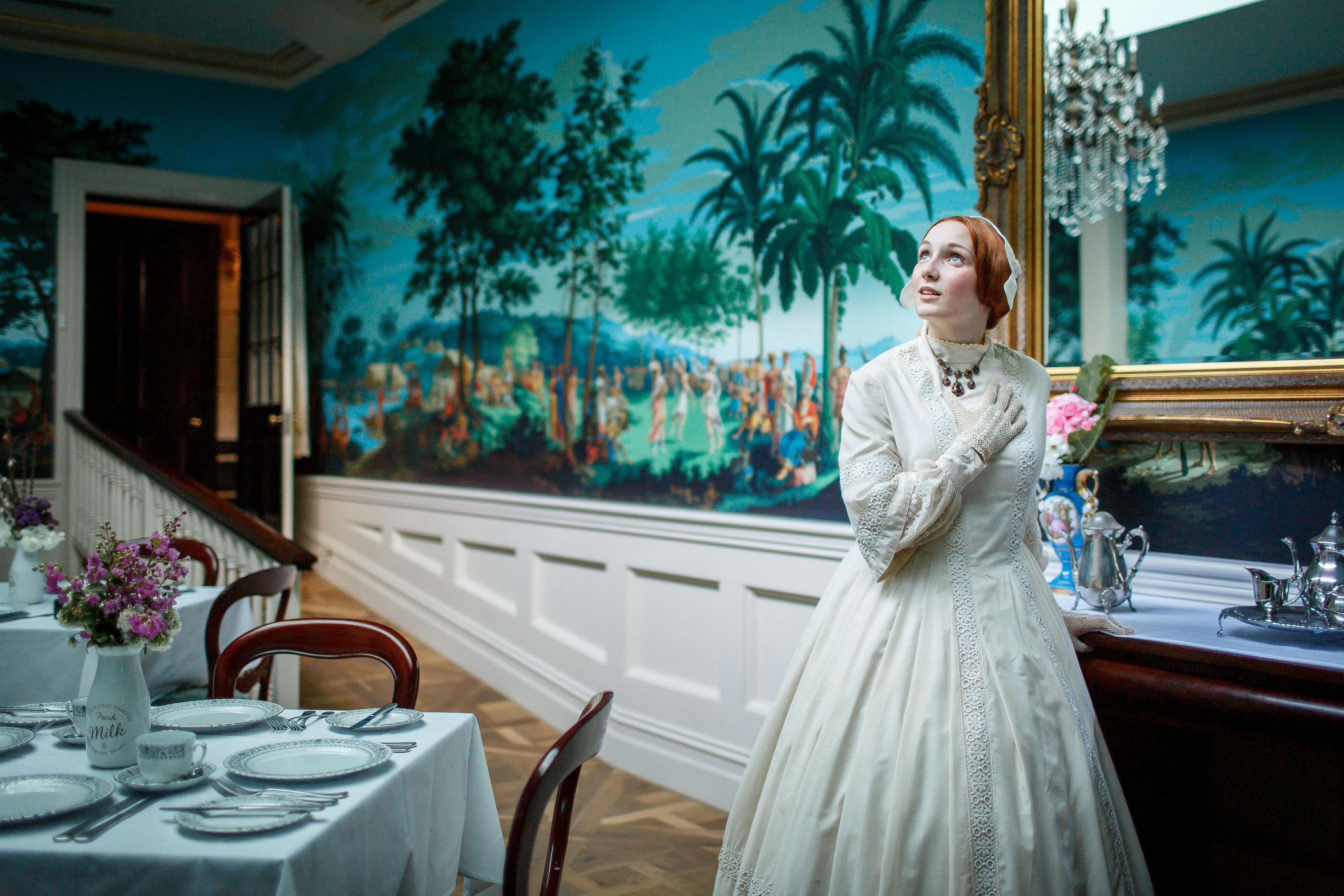A person in a white, vintage gown stands indoors by a table with a silver tea set, beneath a large painting of palm trees.