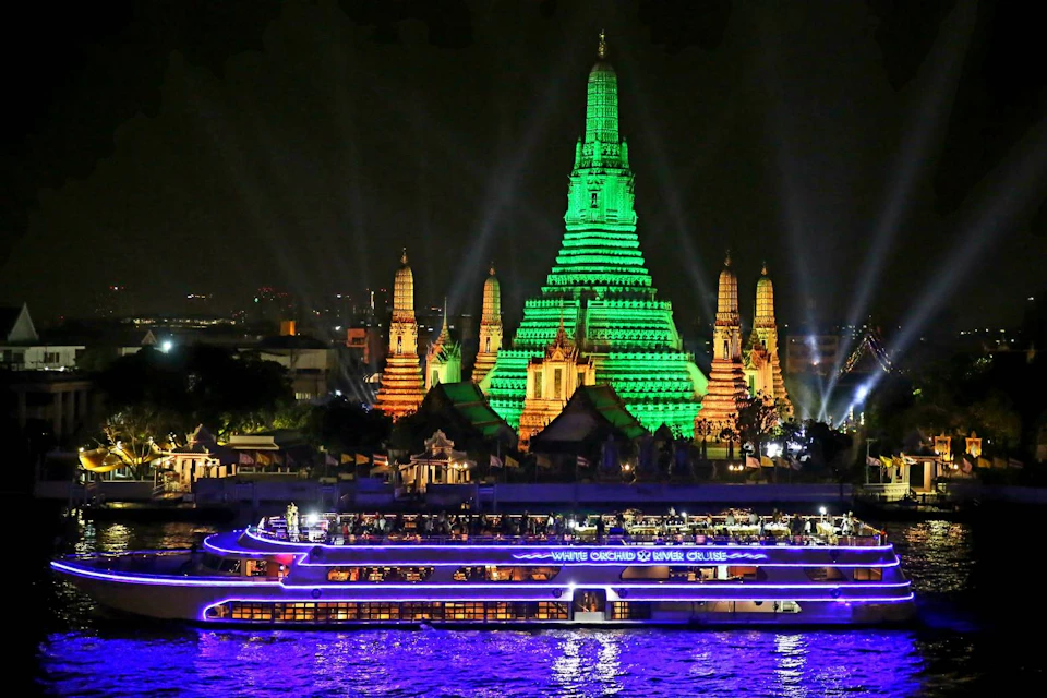 A brightly lit temple with green and orange lights at night, with a river cruise boat in the foreground.