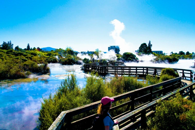 A person wearing a pink cap stands on a wooden walkway overlooking a geothermal area with steam and vegetation under a clear sky.