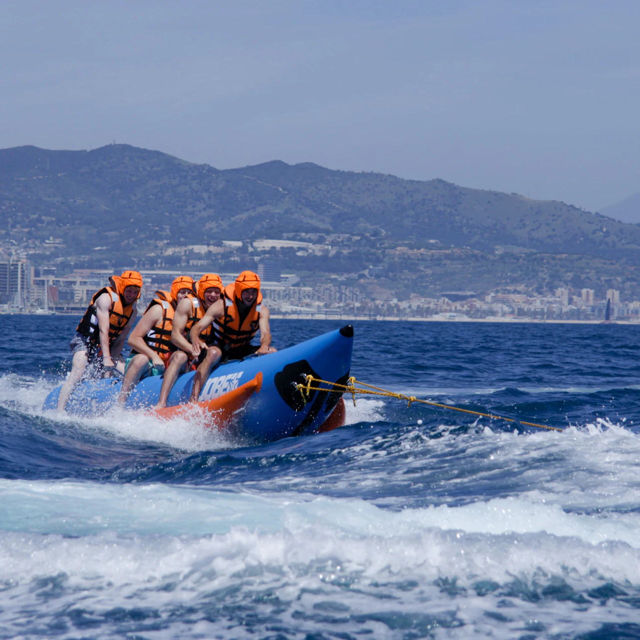 Four people wearing orange helmets and life vests are riding a blue inflatable banana boat on the ocean, with mountains in the background.
